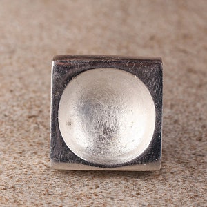 May include: A silver ring with a square band and a round, domed top.