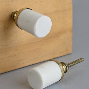 Może przedstawiać: Dwa białe marmurowe gałki do szafek z mosiężną obwódką. Gałki mają cylindryczny kształt i gładkie, polerowane wykończenie.