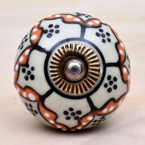 Può includere: Un pomello rotondo in ceramica per mobili con base color crema, decorato con un motivo ripetitivo di linee nere, accenti curvi arancioni e piccoli gruppi di punti neri. Al centro è presente una placca metallica color ottone con una vite.