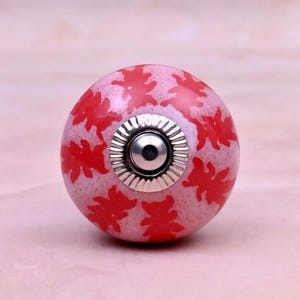 May include: A round ceramic cabinet knob featuring a light pink base with a speckled texture and a repeating pattern of bright red bear silhouettes. The centre of the knob has a circular silver-toned metal hardware fitting with a ridged edge.
