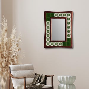 Puede incluir: Un espejo rectangular con un marco de madera marrón oscuro. El marco está acentuado con azulejos verdes y azulejos blancos decorativos con patrones verdes. El espejo está colgado en una pared beige, reflejando la habitación.