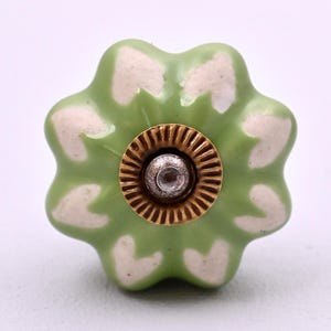 Peut inclure: Un bouton de meuble en céramique en forme de fleur, doté d'un vernis vert clair avec des motifs en forme de cœur blanc sur chaque pétale. Le centre du bouton présente une plaque métallique dorée circulaire avec un bord strié et une vis argentée.