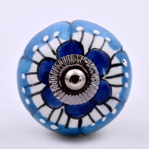 Peut inclure: Un bouton de meuble rond en céramique orné d'un motif floral peint à la main dans des tons bleu et blanc. Le design présente des pétales bleu foncé soulignés de noir sur un fond blanc avec des touches bleu clair et une base en métal argenté.