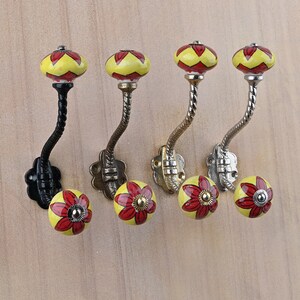May include: Four wall hooks featuring a curved metal stem and two ceramic knobs. Each knob is yellow with red petal patterns. The metal stems vary in finish, including black, antique brass, and polished silver, each with a floral-shaped base plate.