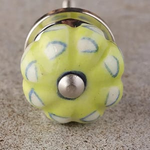 Pode incluir: Um botão de cerâmica amarelo com um padrão de bolinhas brancas e azuis. O botão tem uma base de metal prateado.