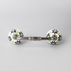May include: A white ceramic drawer pull with black and green floral designs. The pull has a silver metal bar connecting the two knobs.
