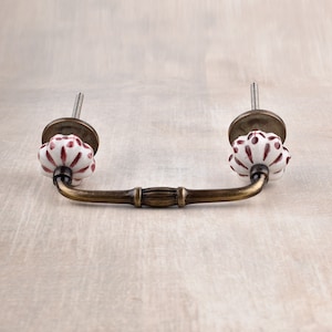 May include: A white ceramic drawer pull with a red floral design and a brass finish. The pull has a curved shape and is attached to a metal base.