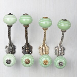 May include: Four wall hooks featuring light green ceramic knobs with white horizontal stripes. Each hook has a twisted metal stem and a decorative floral-shaped base plate. The metal finishes vary in tone, including dark bronze, brass, and silver.