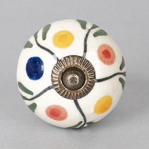 May include: A round, white ceramic cabinet knob with a floral design. The knob features colourful polka dots in yellow, red, and blue, with green leaves and black vines. It has a bronze-coloured metal base.