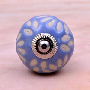 Op de afbeelding: Een ronde keramische meubelknop met een lavendelblauwe glazuurlaag en een herhalend patroon van crèmekleurige, bladvormige uitsparingen. Het midden van de knop is voorzien van een gepolijst zilverkleurig metalen beslag met een geribbelde rand.