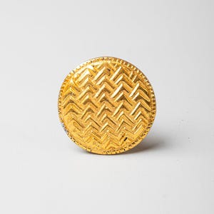 May include: A circular gold-toned button featuring a raised, textured herringbone pattern across its surface. The edge of the button is finished with a fine, notched border detail, creating a distinct rim around the central geometric design.