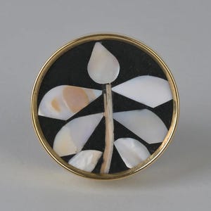 Pode incluir: Um botão redondo com aro dourado, fundo preto e um design de flor estilizado branco feito de madrepérola.