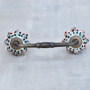 Può includere: Una maniglia per armadio decorativa con una barra in ottone e due pomelli in ceramica. I pomelli sono bianchi con un motivo floreale nero e verde.