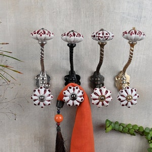 Puede incluir: Cinco ganchos decorativos con pomos de cerámica blanca con diseños florales marrones. Los ganchos son de metal y tienen un diseño retorcido. Los ganchos están montados en una pared gris claro. Una bufanda naranja brillante está colgada en uno de los ganchos.