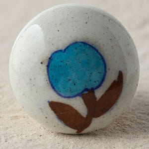 Puede incluir: Pomo de cerámica blanco con un diseño de flor azul. La flor tiene hojas marrones y un centro azul.