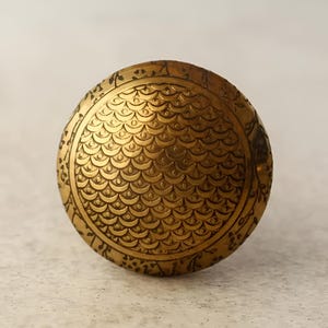 May include: A round, gold-coloured metal knob with a decorative pattern of overlapping circles and a raised, textured centre.