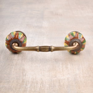 以下が含まれることがあります： 真鍮製のキャビネットプルに、丸いセラミックノブが2つ付いています。ノブは、茶色、黄色、緑の幾何学模様で装飾されています。