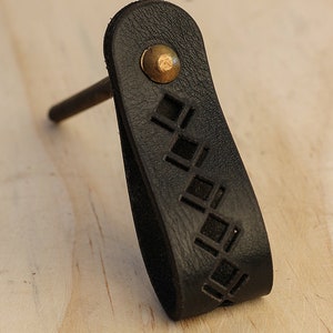 May include: Black leather cabinet pull with a geometric cutout design and a brass knob.