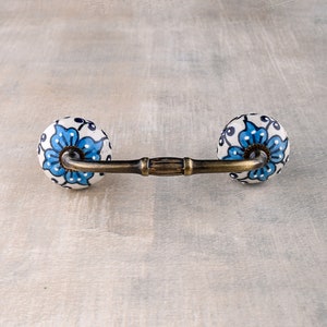 以下が含まれることがあります： 青と白のフローラルデザインのセラミック製の引き出しノブ。ノブは真鍮のバーで2つのセラミックノブを繋いでいます。