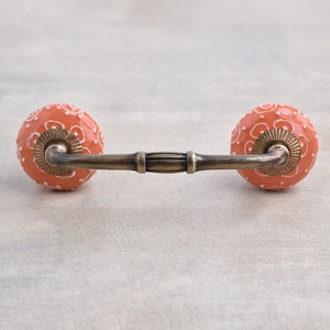 Può includere: Una maniglia per cassetto in ceramica con una base color pesca e un design floreale bianco. La maniglia ha una barra in ottone che collega le due manopole.