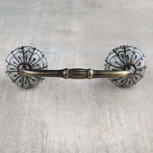 May include: A white ceramic drawer pull with a black floral design and a brass bar connecting the two knobs.
