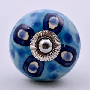 May include: A round ceramic cabinet knob featuring a light blue base with dark blue and white circular patterns. The center of the knob has a silver-colored metal hardware piece with a ridged, circular edge and a smooth, flat center point.