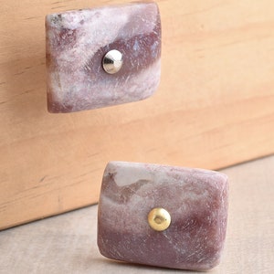 May include: Two square-shaped drawer pulls made of natural stone with a marbled pattern in shades of pink and white. The top pull has a silver knob, while the bottom pull has a gold knob.