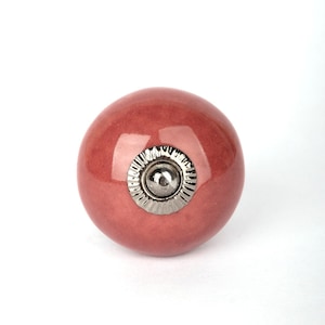 May include: A round, pink ceramic knob with a silver metal accent. The knob is a decorative hardware piece.