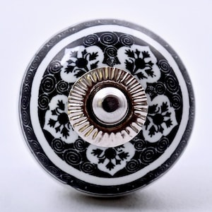 May include: A round ceramic cabinet knob featuring a black and white floral pattern with swirling details. The center of the knob has a polished silver-colored metal hardware piece with a fluted edge and a smooth, raised circular dome in the middle.
