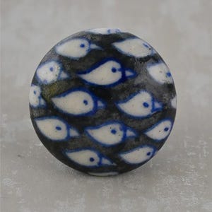 Op de afbeelding: Ronde decoratieve meubelknopp met een patroon van witte vogels met blauwe details op een donkere achtergrond. De knop, waarschijnlijk van keramiek, is geschikt voor meubels of kastdeuren. Diameter ongeveer 3 cm.