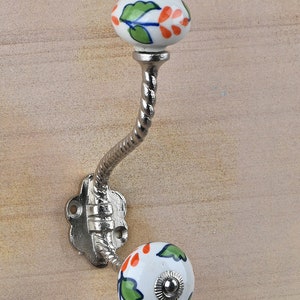 Può includere: Un gancio argentato con uno stelo attorcigliato e due pomelli in ceramica bianca. I pomelli sono decorati con foglie verdi e fiori arancioni.