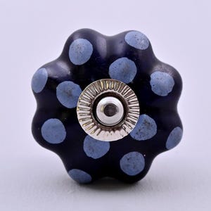 May include: A dark blue ceramic drawer knob with a scalloped, flower-like shape and light blue polka dots. The center features a circular, silver-toned metal hardware piece with a ridged, sunburst-style edge and a smooth, reflective dome.