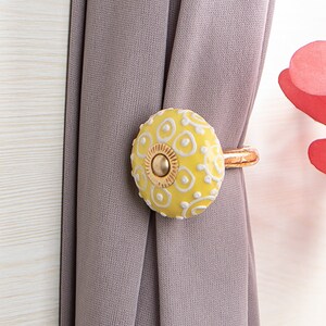 May include: Yellow ceramic curtain tieback with a white floral design and a gold metal ring.