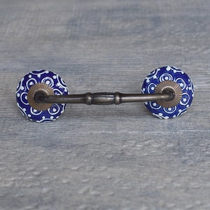 Op de afbeelding: Een blauwe en witte keramische ladegreep met een messing stang die de twee knoppen verbindt. De knoppen hebben een cirkelvormig ontwerp met witte stippen op een blauwe achtergrond.