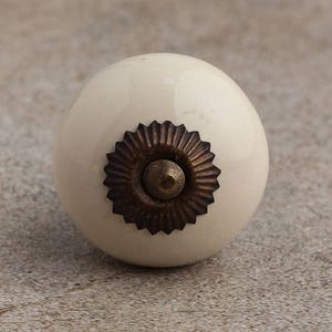 Peut inclure: Un bouton de meuble rond en céramique blanc cassé avec une finition lisse et brillante. Le centre du bouton est orné d'une plaque métallique de couleur bronze antique foncé, dotée d'un bord festonné en forme de soleil et d'un petit clou central.