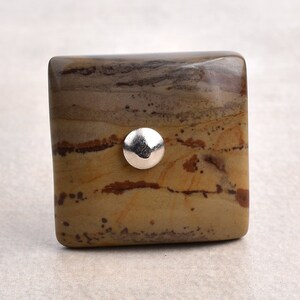 Pode incluir: Botão quadrado para armário feito de pedra com padrão marrom e bege com um detalhe em metal prateado.