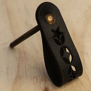 May include: A black leather cabinet pull with a star and circle cut-out design. The pull has a brass screw and a brass accent on the top.