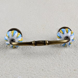 May include: A brass cabinet pull with two white ceramic knobs. The knobs have a blue and yellow striped pattern.