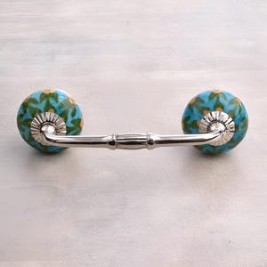 May include: A silver drawer pull with two blue and green ceramic knobs with a floral pattern. The knobs have a gold trim.