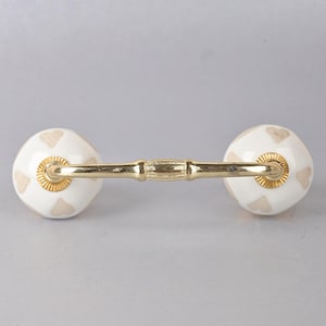 May include: White ceramic drawer pull with gold accents and a heart pattern. The pull has a gold metal bar connecting the two knobs.
