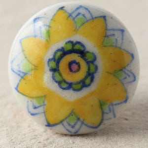 Könnte beinhalten: Ein weißer Keramik-Knopf mit einem gelben Blumendesign. Die Blume hat einen blauen Umriss und eine grüne Mitte.