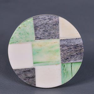 Puede incluir: Posavasos redondo hecho de hueso e incrustaciones de piedra. El posavasos tiene un patrón de tablero de ajedrez en blanco, verde y gris.