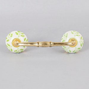 May include: A gold metal drawer pull with two white ceramic knobs. The knobs have a green floral design.