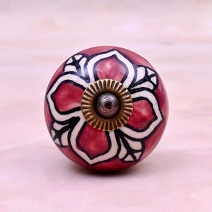 May include: A round ceramic cabinet knob featuring a deep red base colour with a prominent white and black floral pattern. The centre of the knob is accented with a circular, ridged brass-toned metal hardware piece with a small dark centre point.