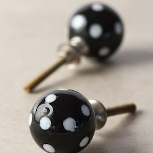 Puede incluir: Dos pomos de armario negros con lunares blancos. Los pomos son redondos con un acabado brillante y lunares blancos. Cada pomo tiene un vástago de metal y una arandela de vidrio o plástico transparente.