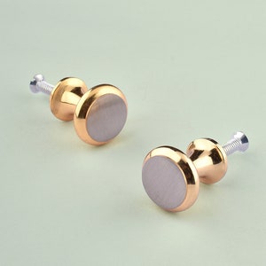 May include: Two gold-toned cabinet knobs with a brushed silver finish. Each knob has a round base and a raised, circular top.