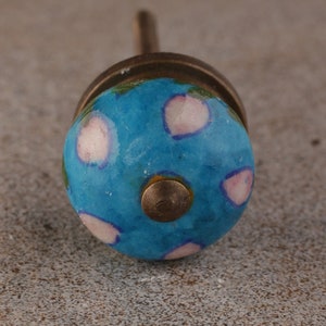 May include: A close-up of a decorative cabinet knob. The knob features a round, blue ceramic design with white and pink accents. The base and stem are a bronze color.