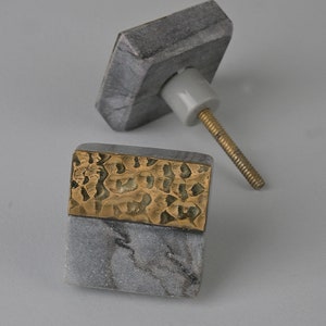 May include: Two square cabinet knobs made of grey marble with a gold metal accent. One knob has a smooth surface and the other has a hammered metal accent.