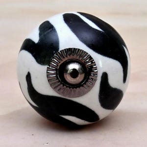 May include: A spherical ceramic cabinet knob featuring a high-contrast black and white abstract pattern. The center of the knob is adorned with a circular, metallic silver-toned hardware piece that has a ridged, scalloped edge and a smooth dome.