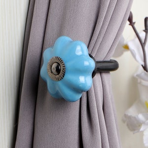May include: A blue ceramic curtain tieback with a silver knob and a black metal hook. The tieback is attached to a gray curtain.